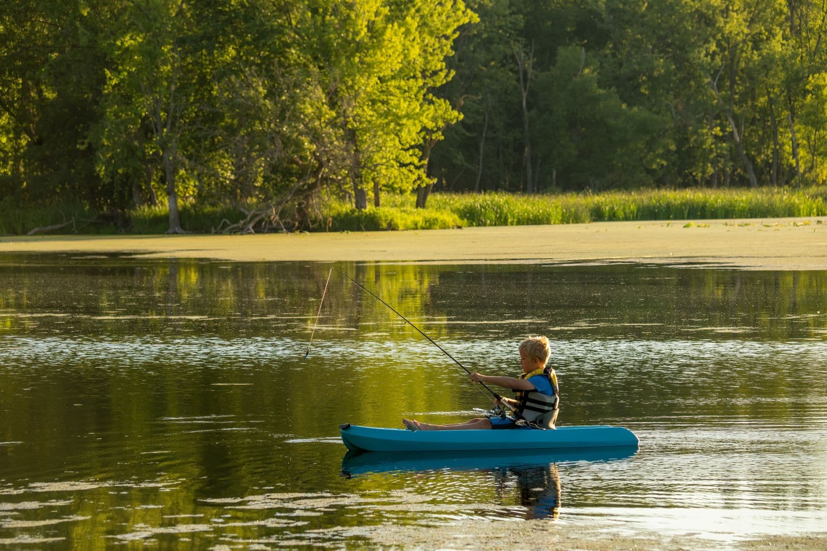 A child in a life jacket fishing from a kayak on a calm, tree-lined lake